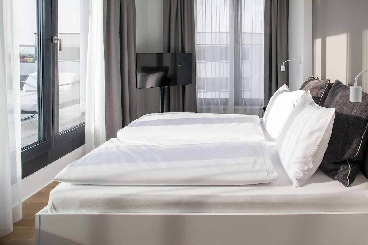 Una habitación de hotel moderna con una cama grande, un televisor de pantalla plana montado en la pared y vistas de la ciudad a través de amplias ventanas.