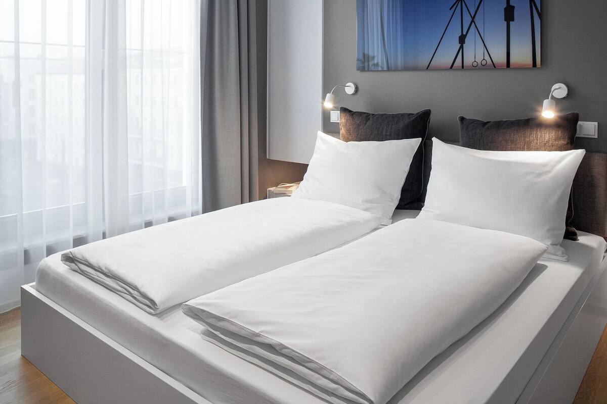 Una cama doble con sábanas blancas, una mesita de noche con lámpara y un cuadro en la pared.