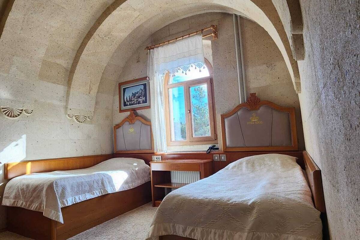 A room with two beds, a wooden headboard, a window with curtains, and a wall-mounted TV.