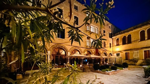 A courtyard with outdoor seating, a building with arched windows, and a tree in the foreground. - Domus Ciliota (Venice)