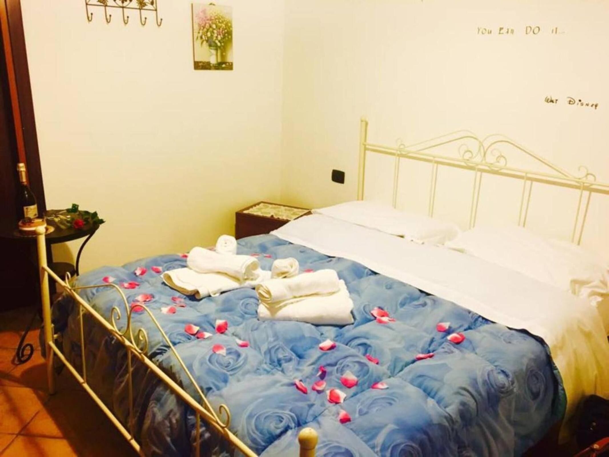 A neatly made bed with blue floral bedding and white pillows, a small bedside table with a bottle and a flower, and a wall-mounted hook with a picture.