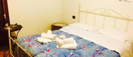 A neatly made bed with blue floral bedding and white pillows, a small bedside table with a bottle and a flower, and a wall-mounted hook with a picture.