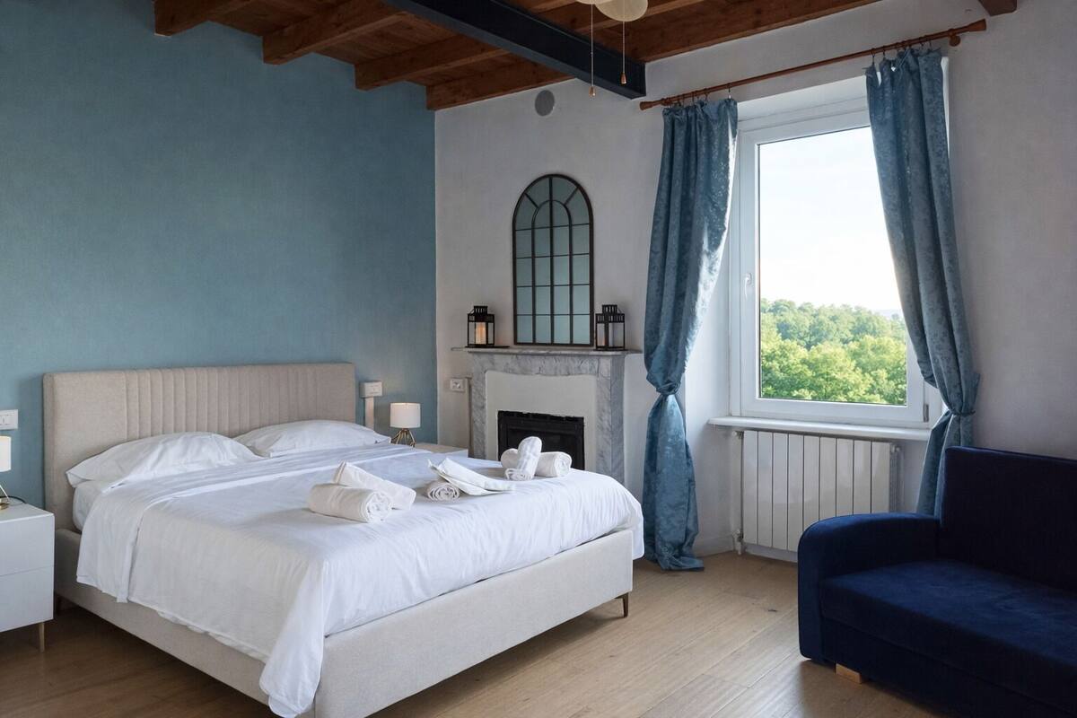 Een slaapkamer met een bed, een nachtkastje, een blauwe fauteuil, een open haard en een raam met gordijnen.
