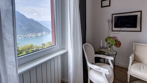 A room with a view of a lake and mountains, a white chair, a small table with a vase of flowers, and a TV on the wall. - Essentia Guest House  (Faggeto Lario)