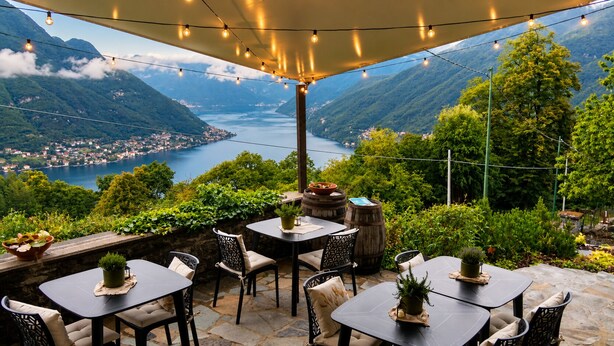 An outdoor patio with tables and chairs, surrounded by greenery and overlooking a valley with a lake and mountains. - Essentia Guest House  (Faggeto Lario)