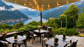 Un patio exterior con mesas y sillas, rodeado de vegetación y con vistas a un valle con un lago y montañas.
