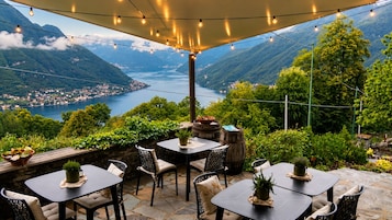 An outdoor patio with tables and chairs, surrounded by greenery and overlooking a valley with a lake and mountains.