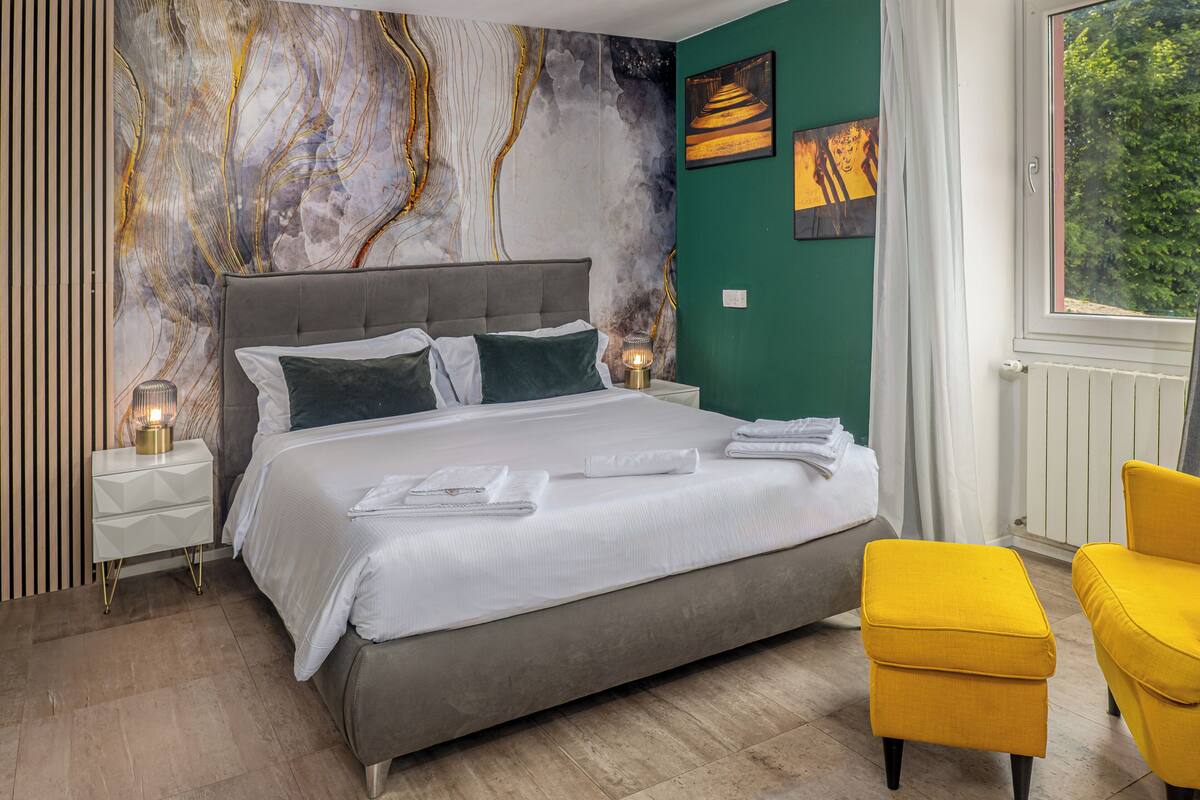 Een moderne slaapkamer met een groot bed, een nachtkastje met een lamp, een geel poufje en een raam met uitzicht op groen.