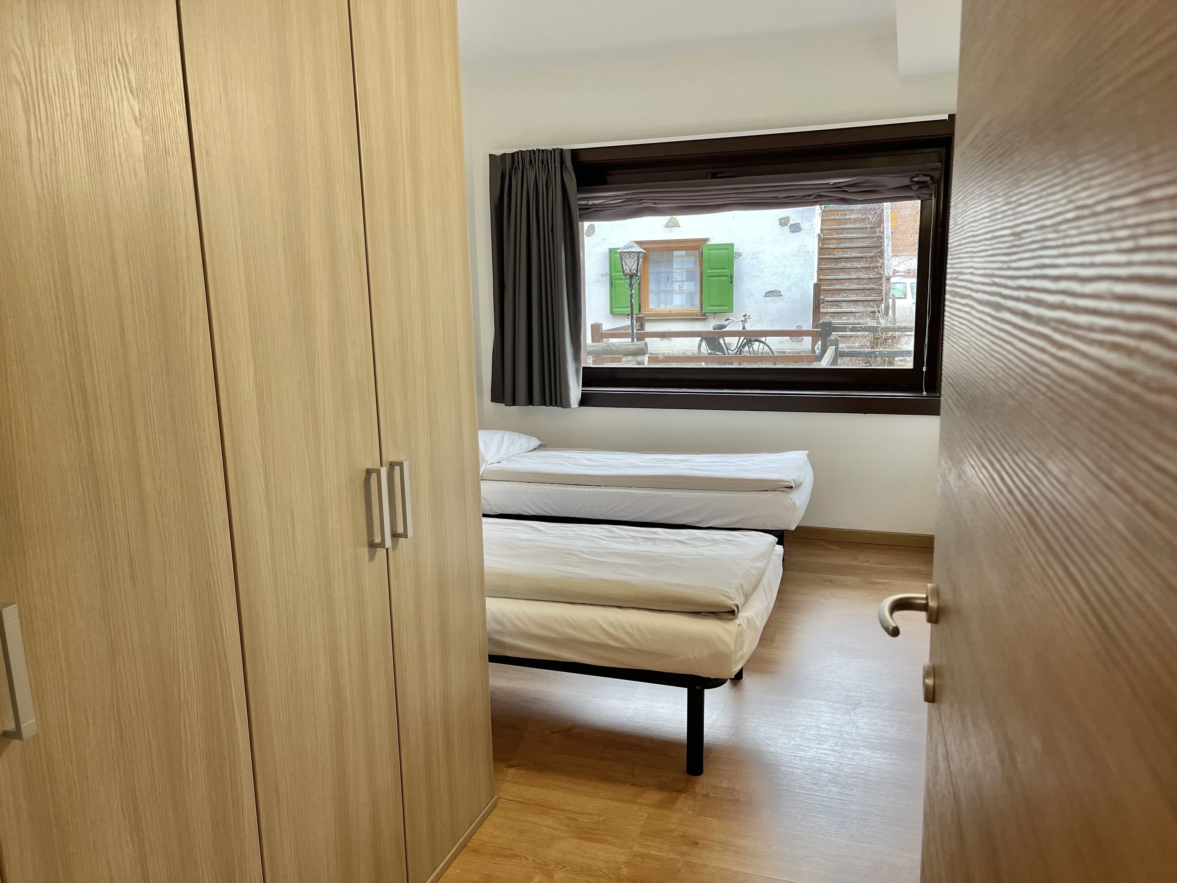 A room with a wooden wardrobe, two single beds, a window with curtains, and a view of the outside.