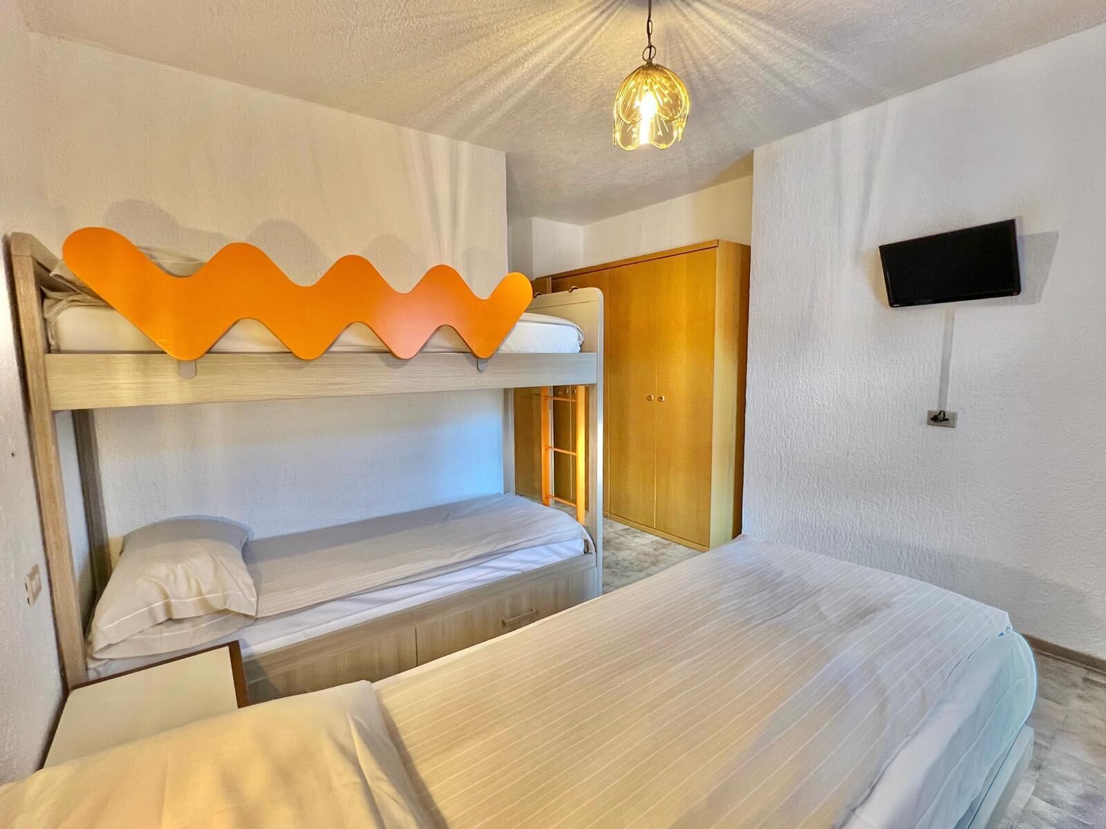 A bunk bed room with a wooden wardrobe, a television mounted on the wall, and a hanging light fixture.