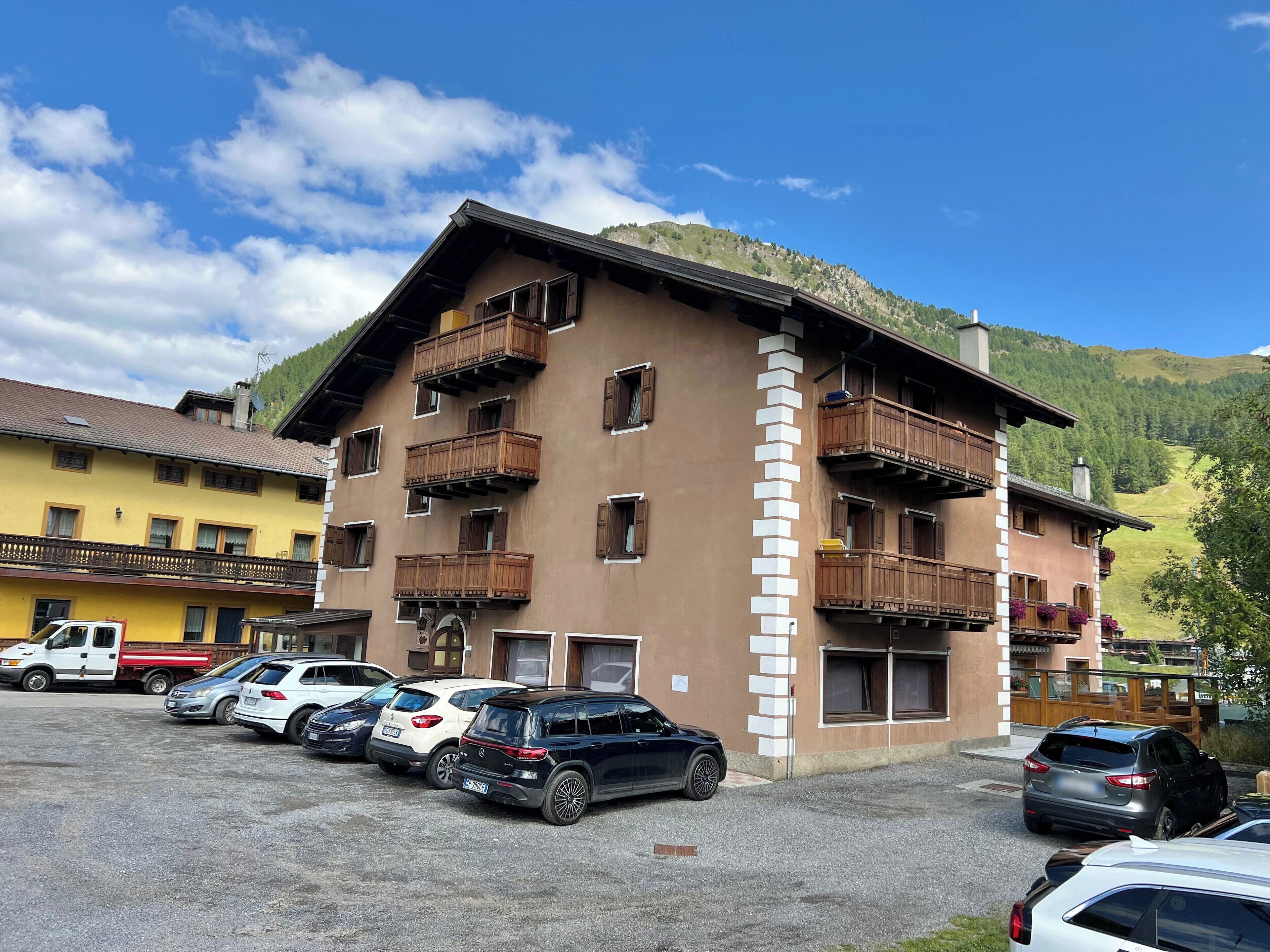 Un immeuble à plusieurs étages doté de balcons en bois, un parking où sont garées plusieurs voitures, et un paysage montagneux en arrière-plan.