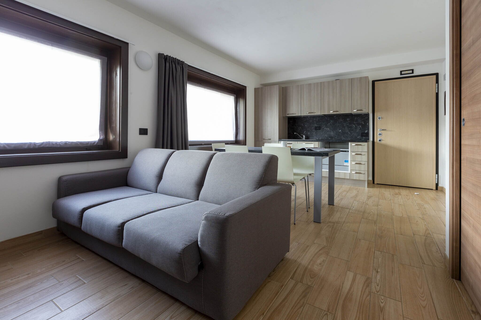 A modern living room with a grey sofa, a dining area with a white table and chairs, a kitchen with wooden cabinets, and a large window with curtains.