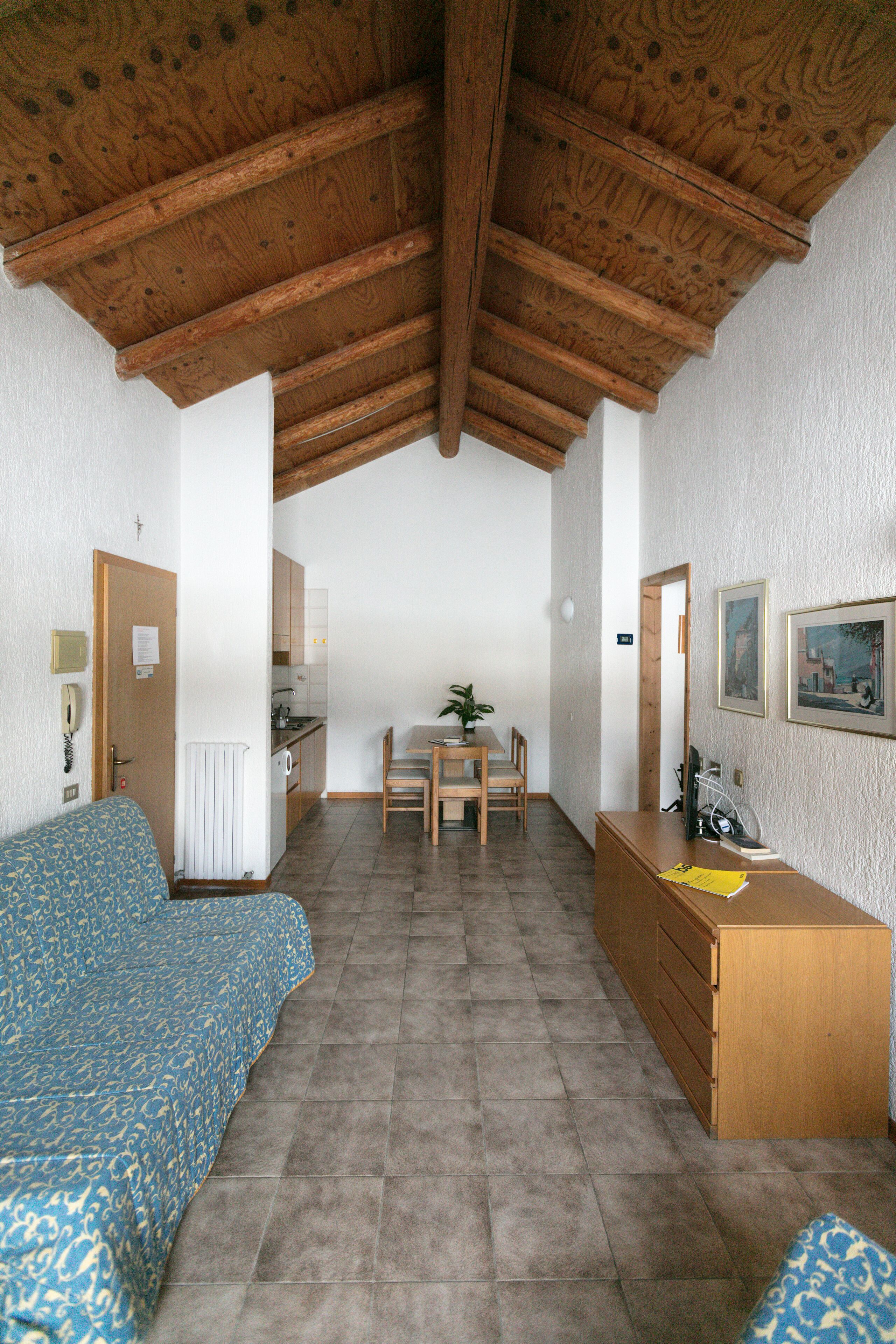A room with a wooden ceiling, a bed, a desk, a chair, a radiator, and a small kitchen area.