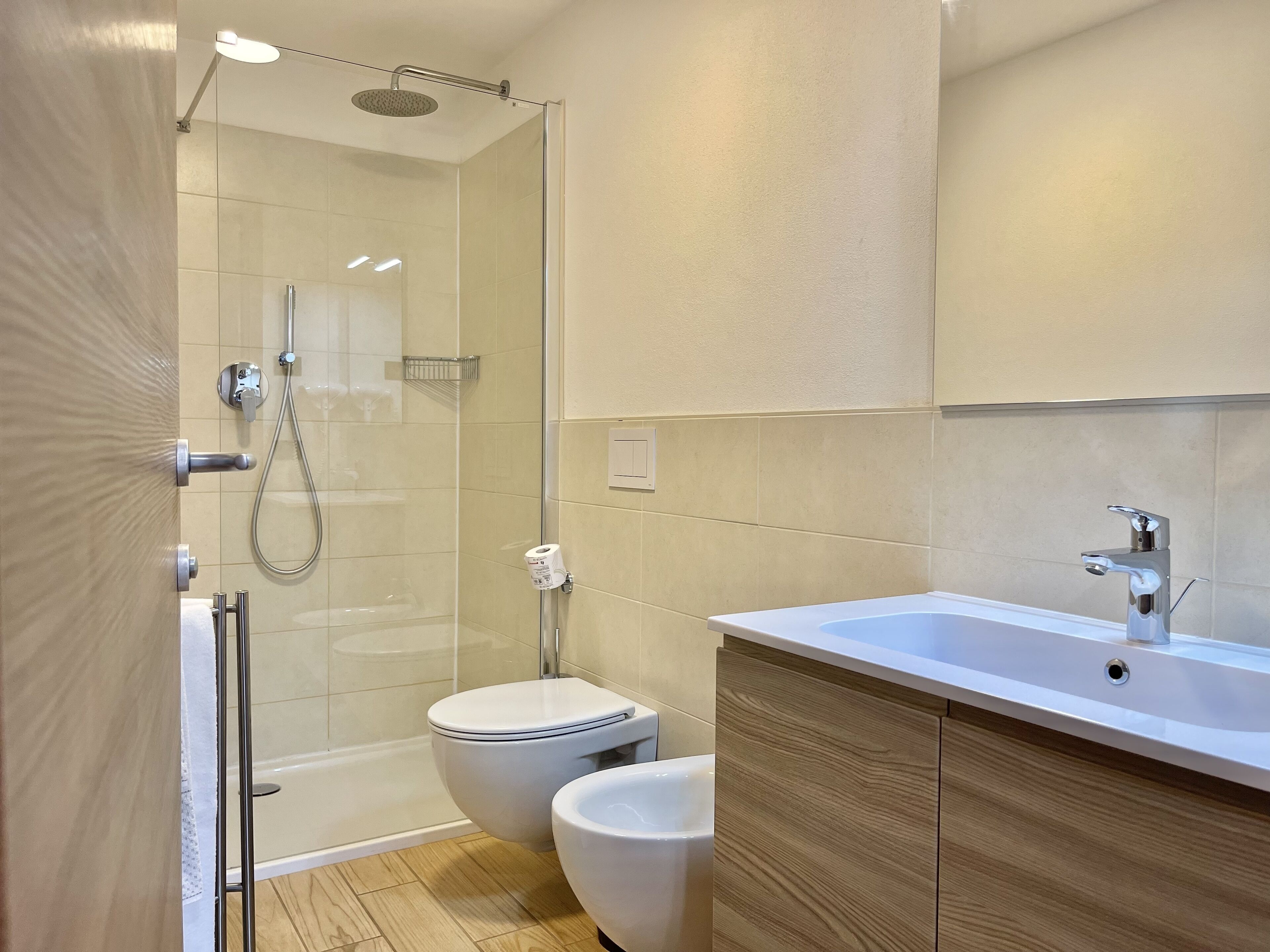 A modern bathroom with a glass shower enclosure, a toilet, a bidet, and a sink with a wooden cabinet.