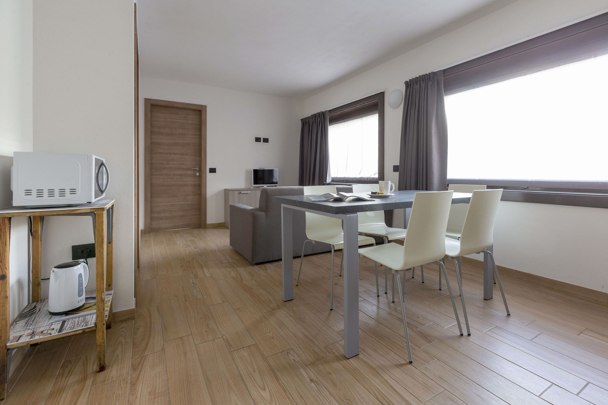 A modern, well-lit room with a wooden floor, a dining table with chairs, a microwave, a kettle, and a television on a small cabinet.