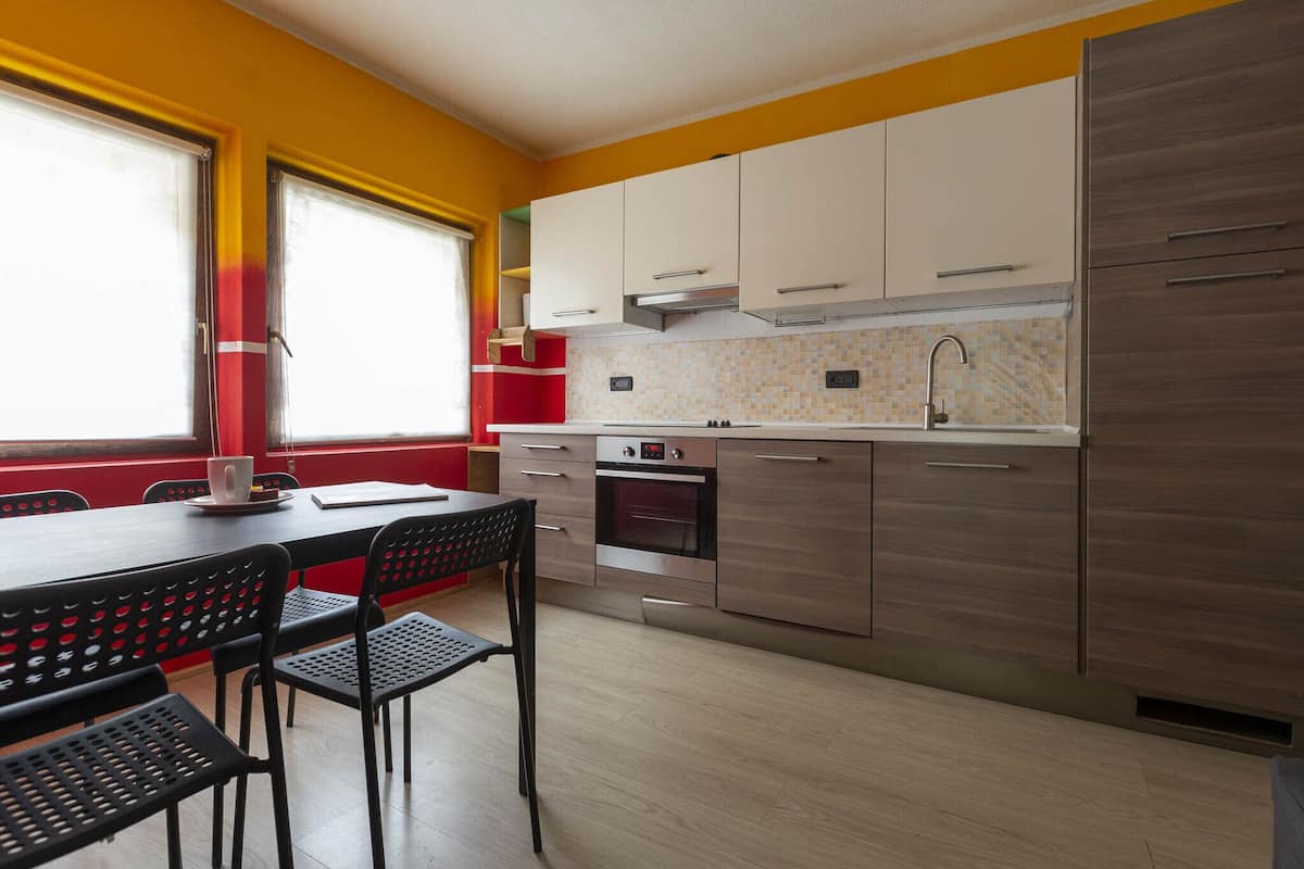Une cuisine moderne avec des meubles en bois, un four encastré et un coin repas comprenant des chaises noires et une table rouge.