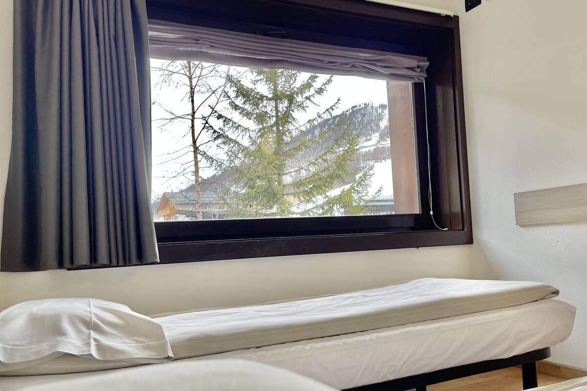 Un lit simple avec un matelas et un oreiller blancs, une fenêtre donnant sur des arbres et des immeubles, et un rideau à moitié tiré.