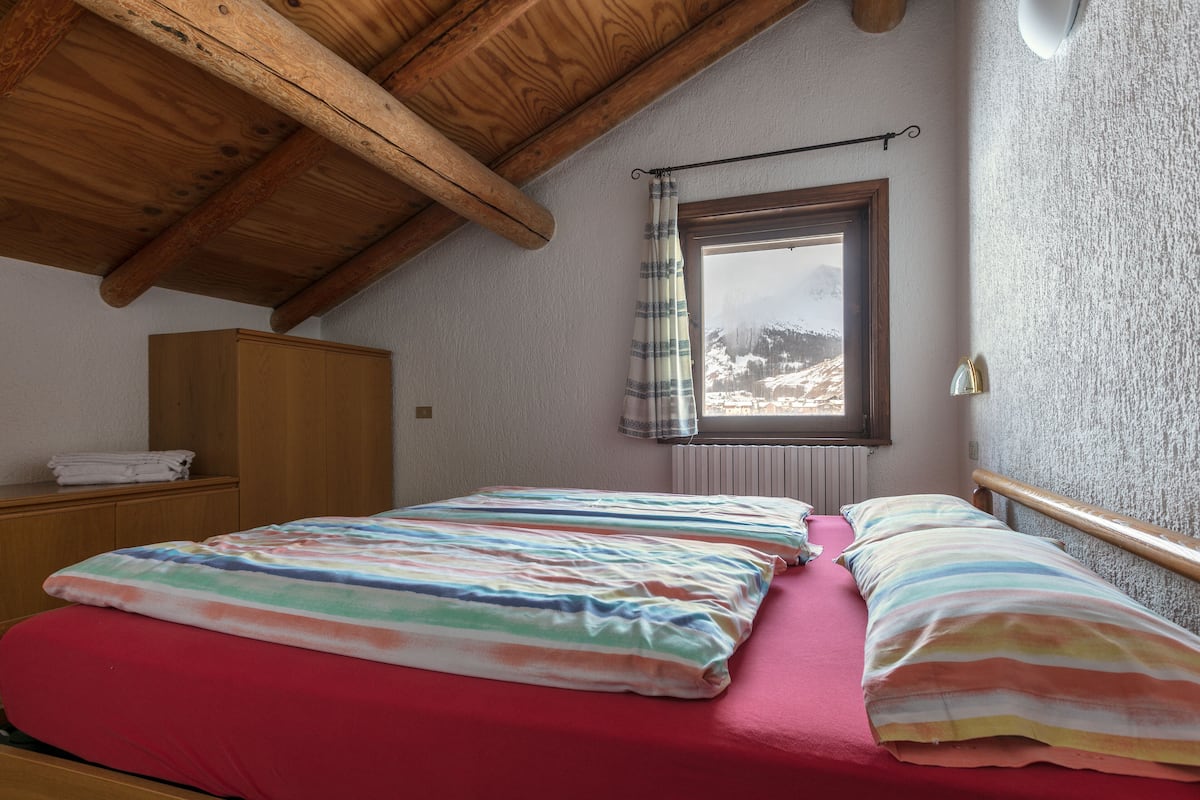 Une chambre avec un plafond en bois, une fenêtre donnant sur des montagnes enneigées, un lit avec une literie à rayures et une armoire en bois.