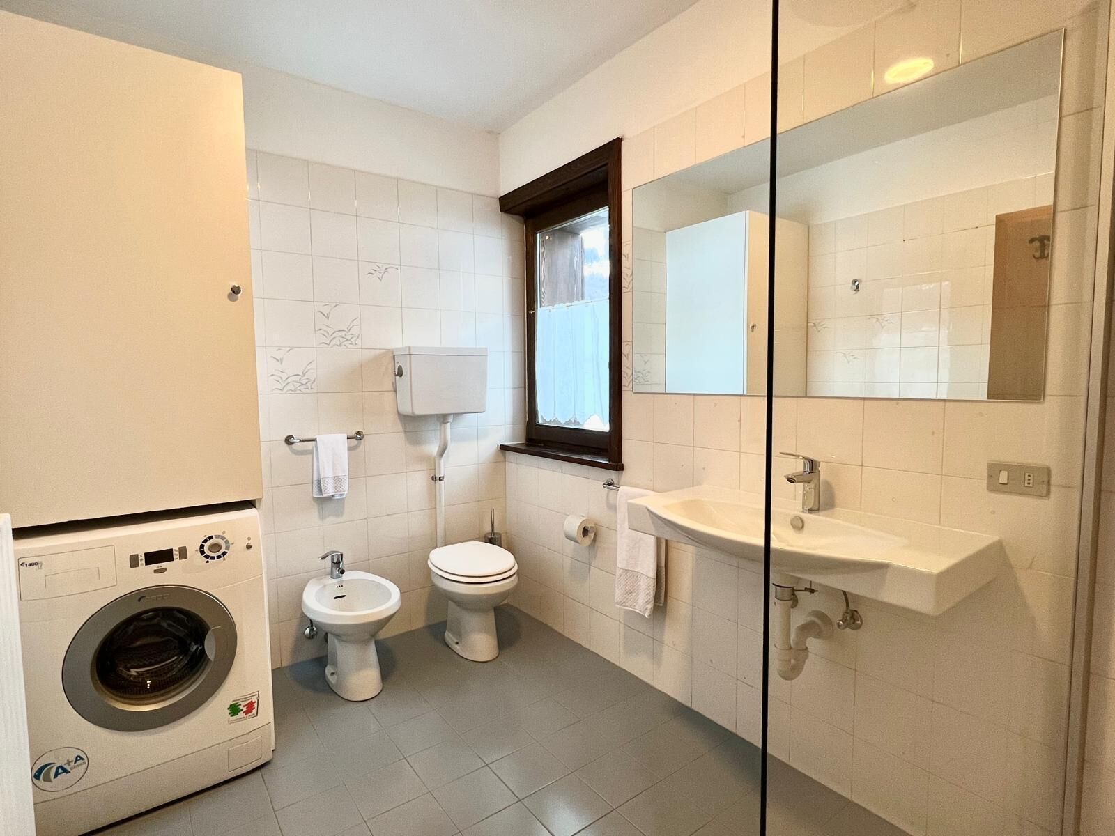 A compact bathroom with a washing machine, two toilets, a sink, and a window with a view.
