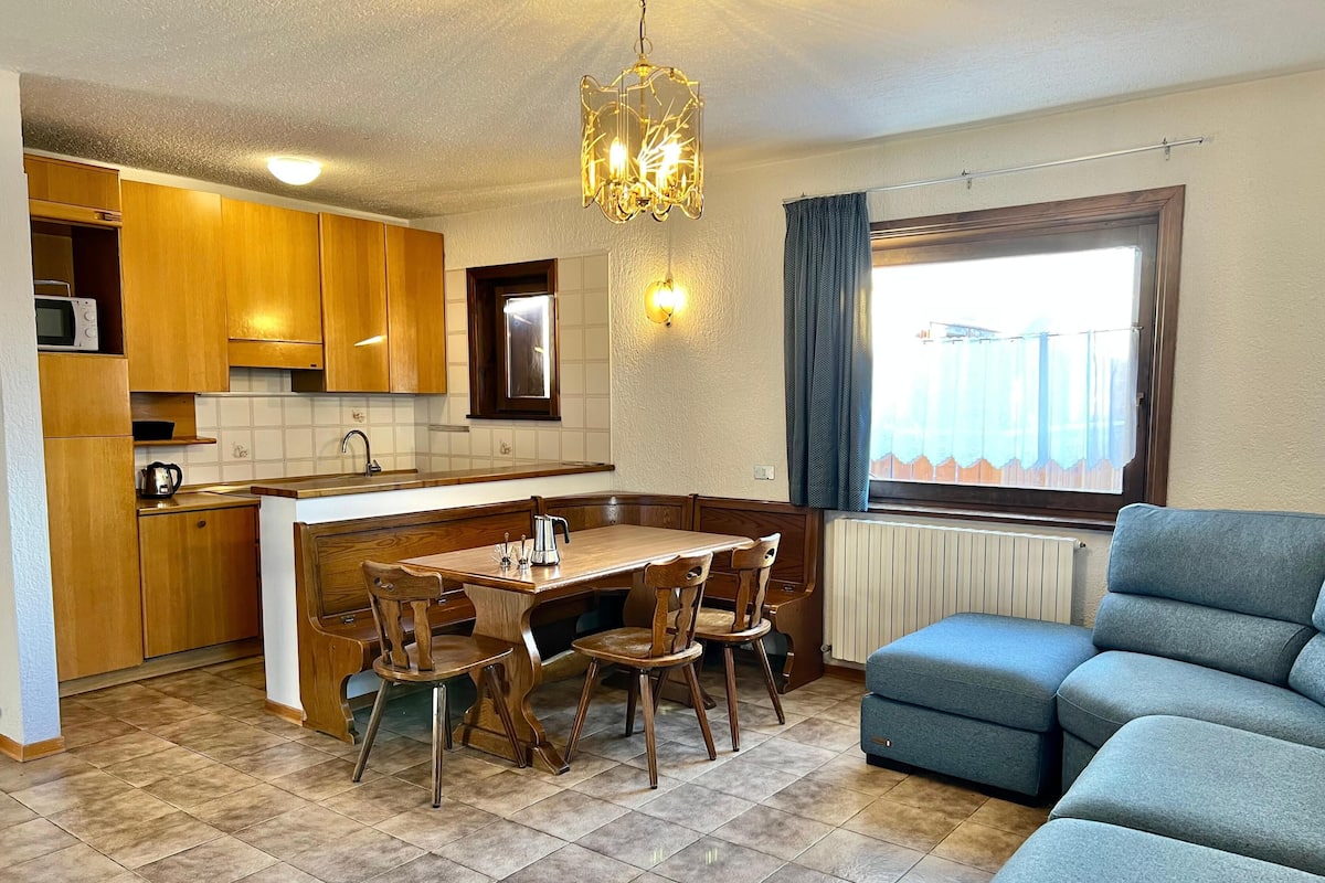 Une cuisine de taille réduite avec des meubles en bois, une table à manger avec des chaises, un lustre et une fenêtre avec des rideaux.