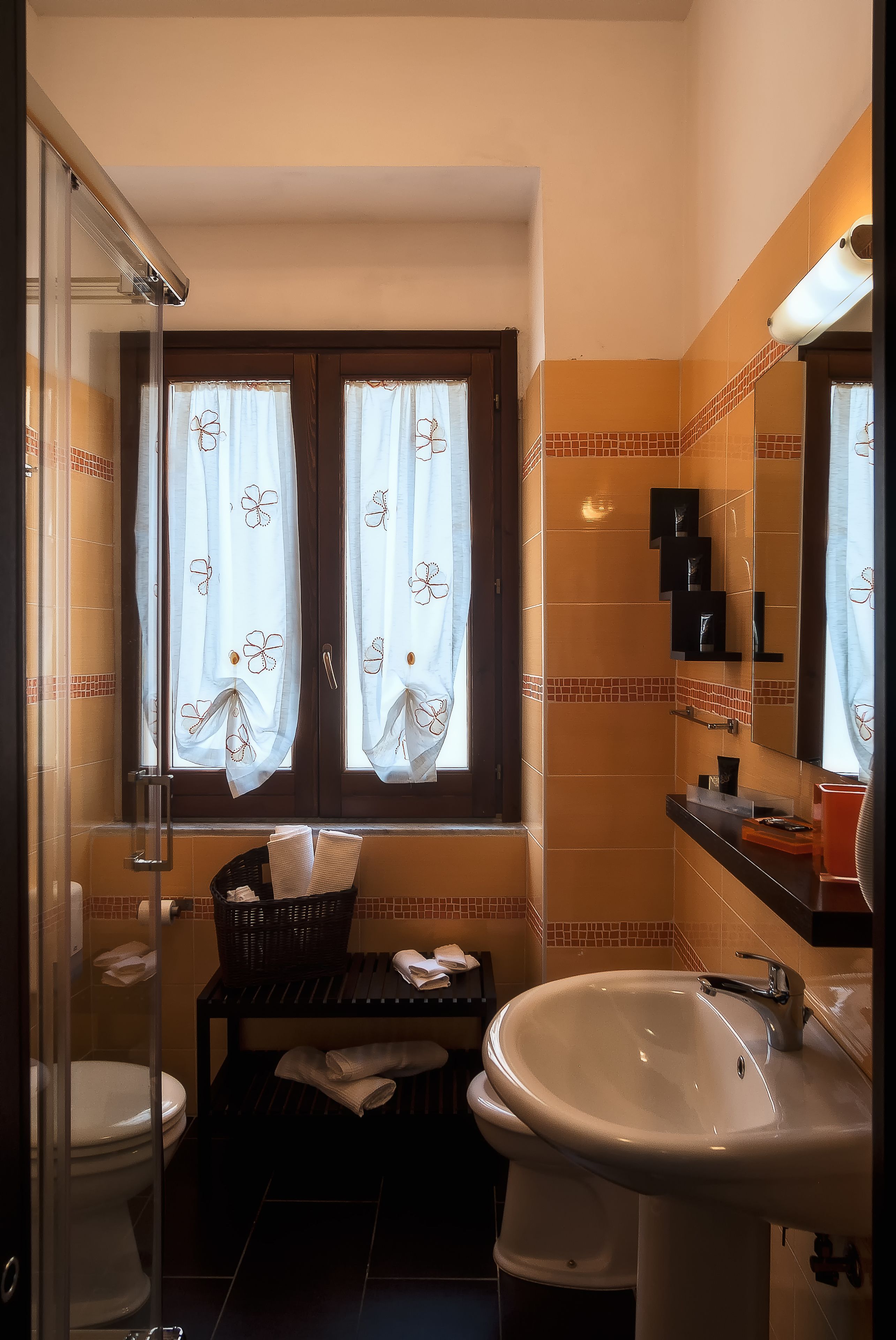 Een badkamer met toilet, wastafel en douchecabine. Er is een raam met gordijnen, een spiegel en een plank met toiletartikelen.