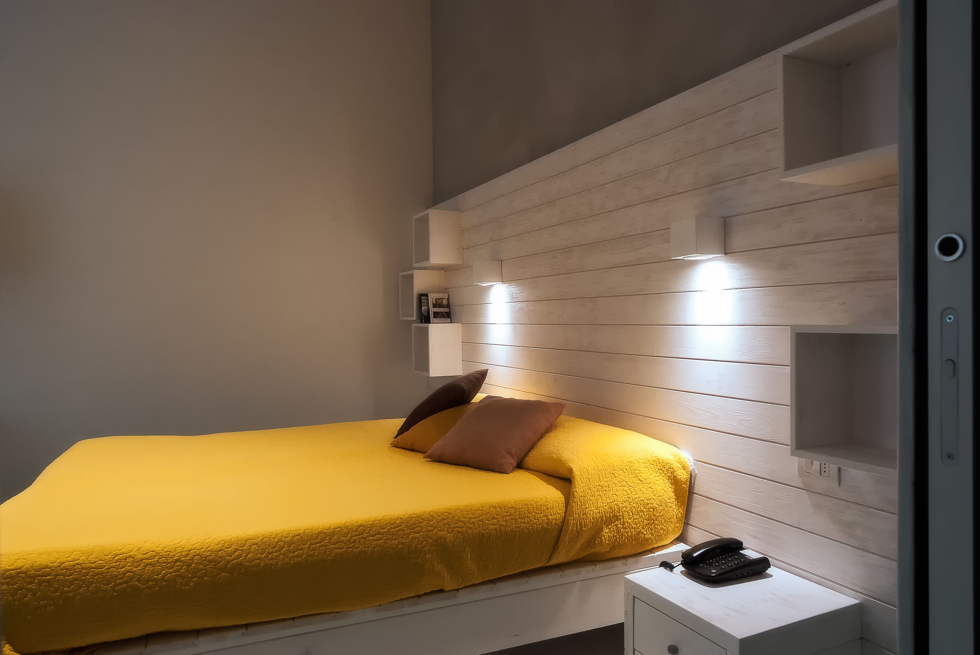 A bed with a yellow bedspread and two pillows, a bedside table with a phone, and built-in shelves with lighting.