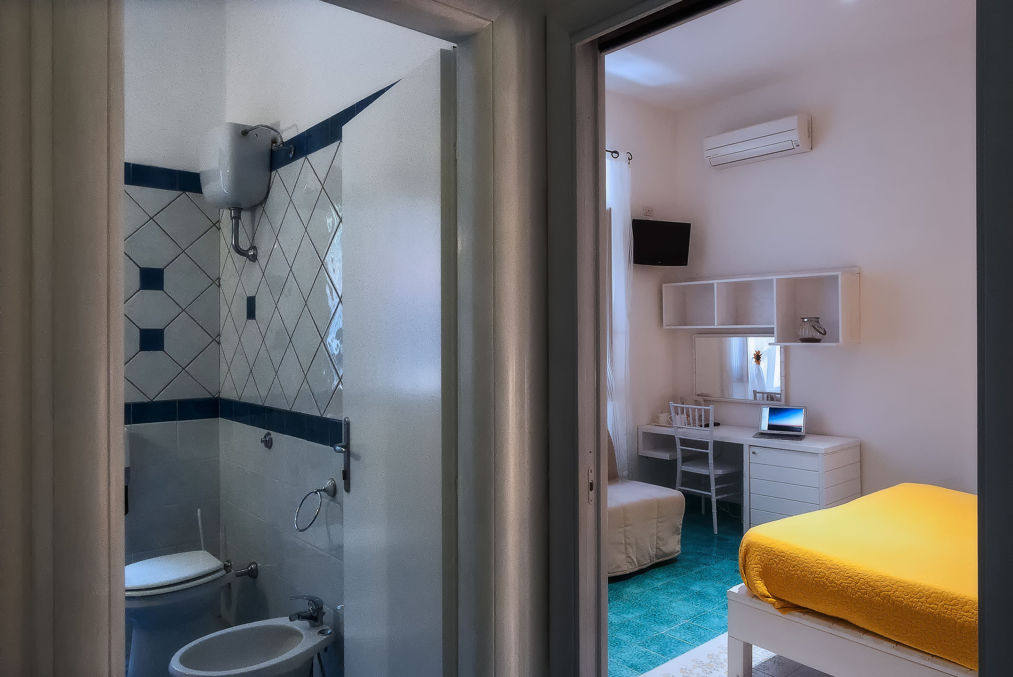A bathroom with a toilet, sink, and shower. There is a bed with a yellow blanket and a desk with a computer.