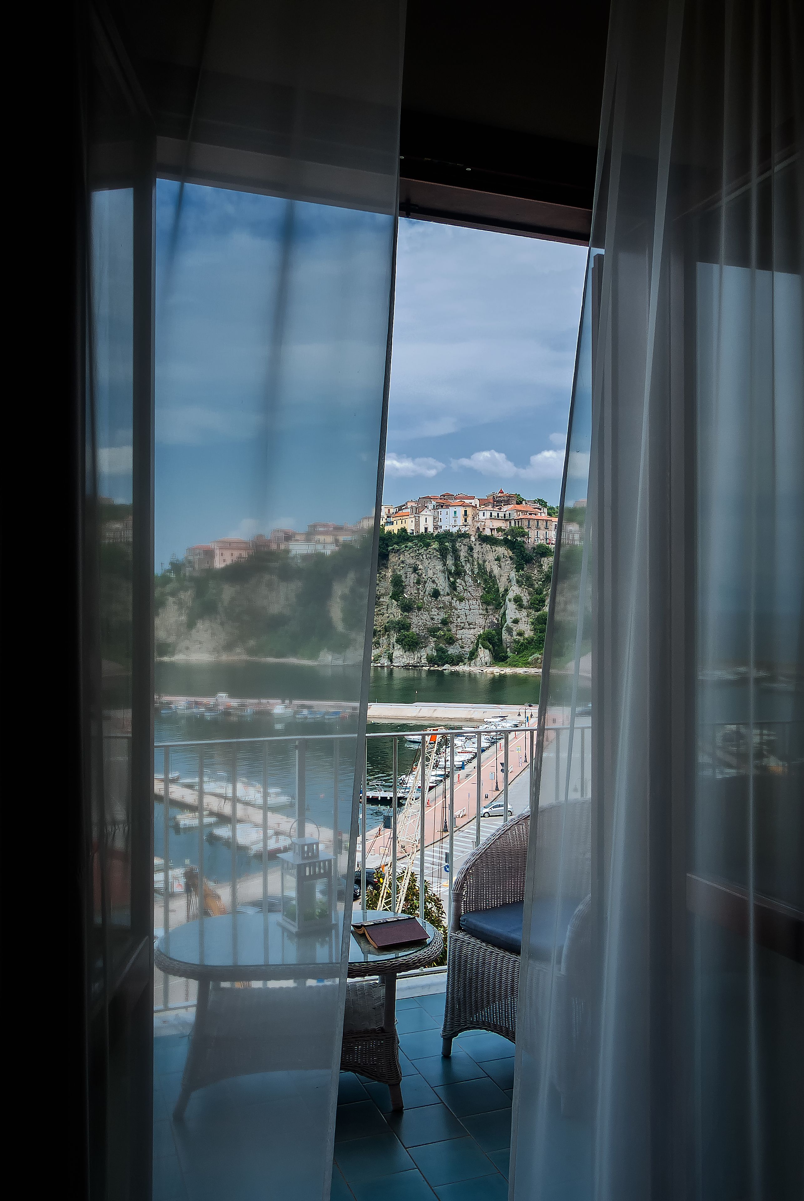 A balcony with a view of a river, buildings on a cliff, and a wicker chair.