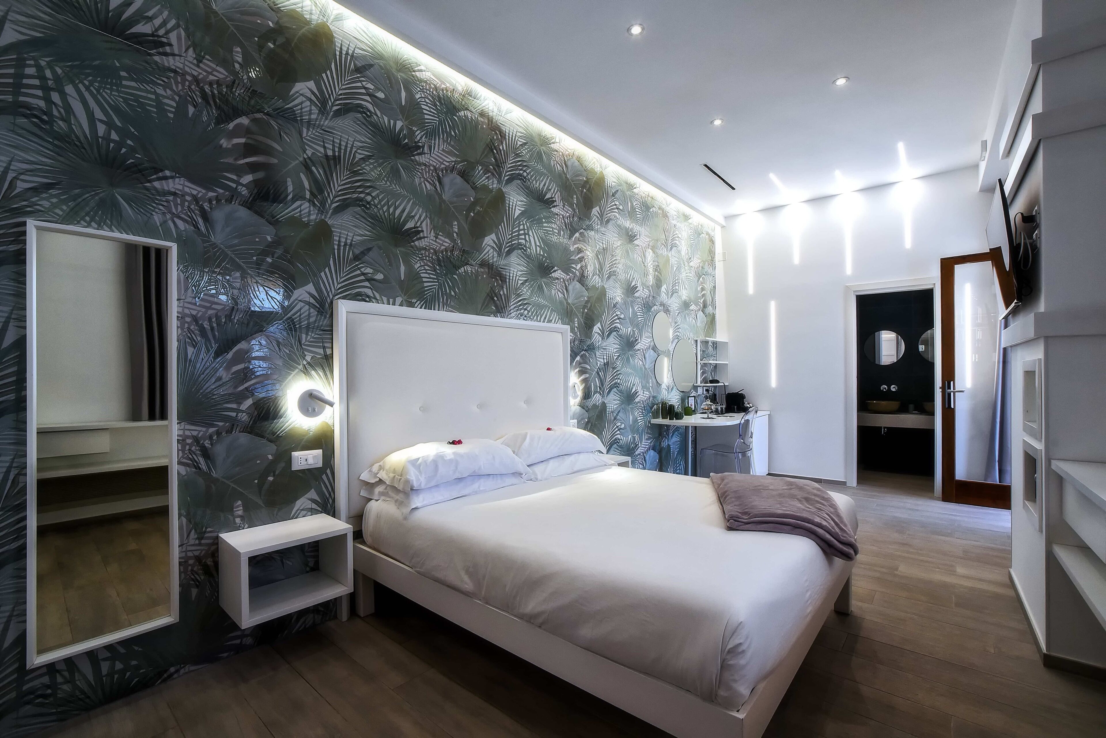 A modern bedroom with a large bed, a bedside table, a mirror, and a wall with a tropical leaf pattern.