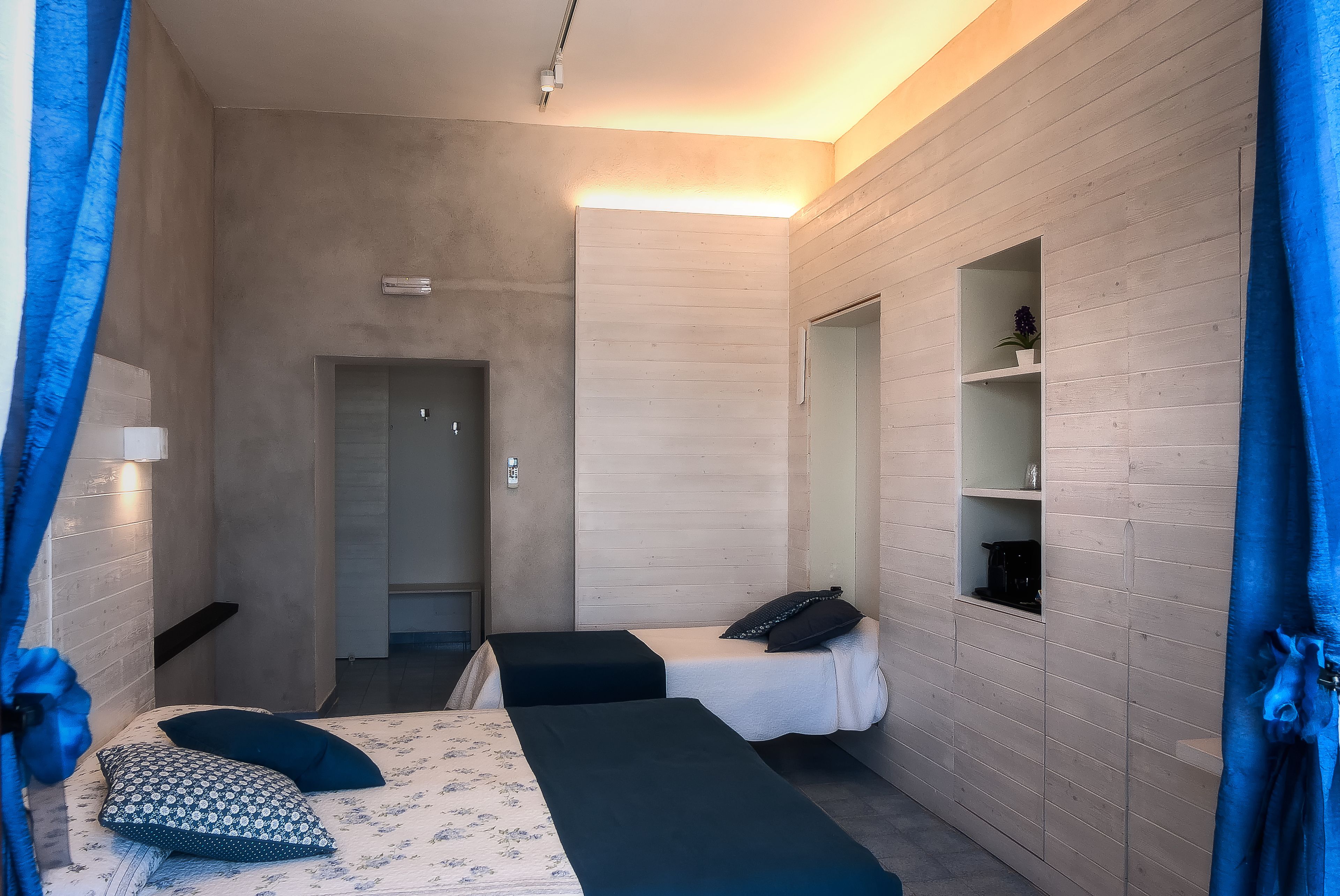 Una habitación moderna con dos camas, un pequeño estante con una planta y una puerta que da a otra habitación.