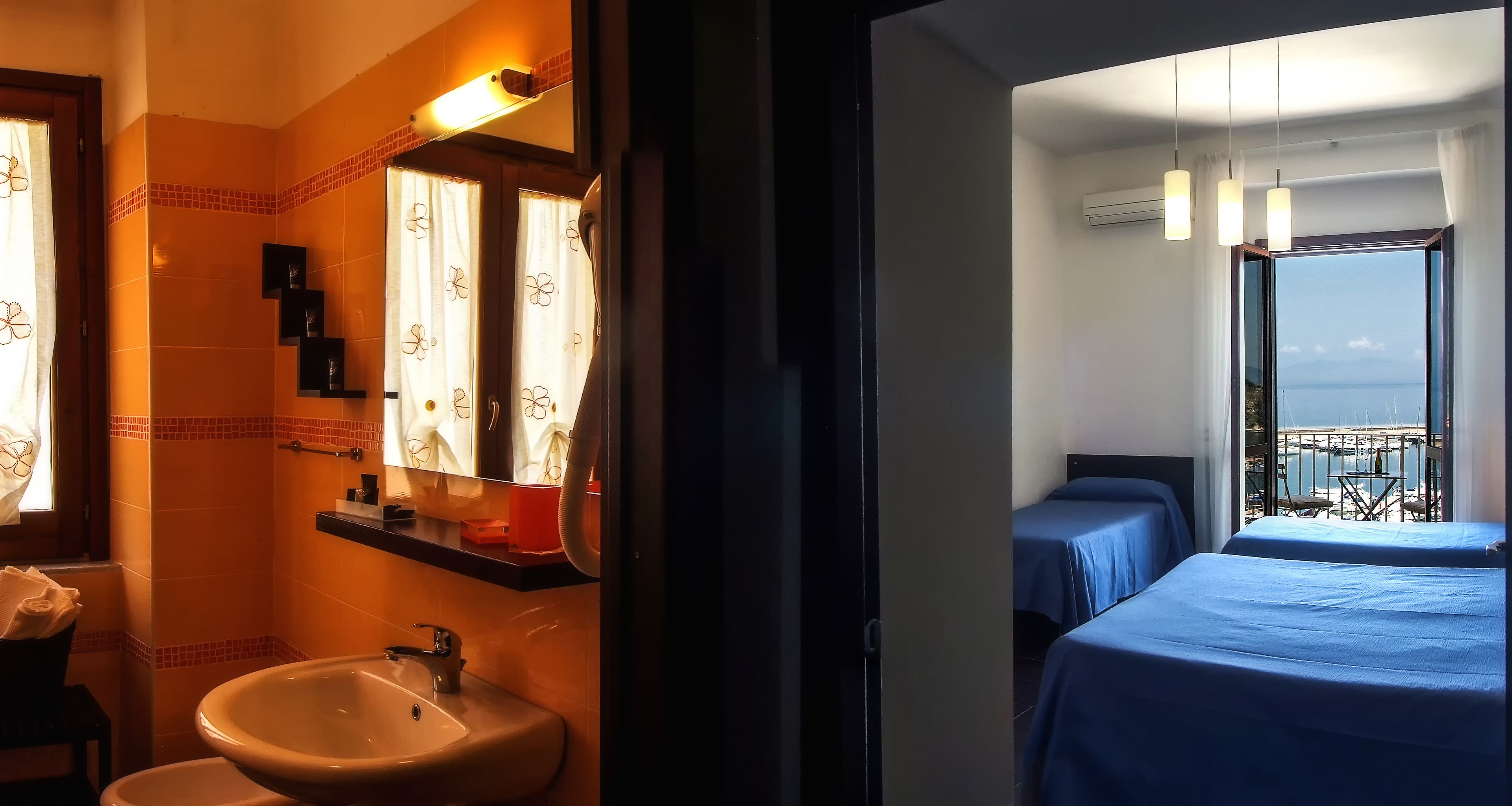 A hotel room with a bathroom featuring a sink, mirror, and towel rack. The room has two beds with blue bedding and a balcony with a view.