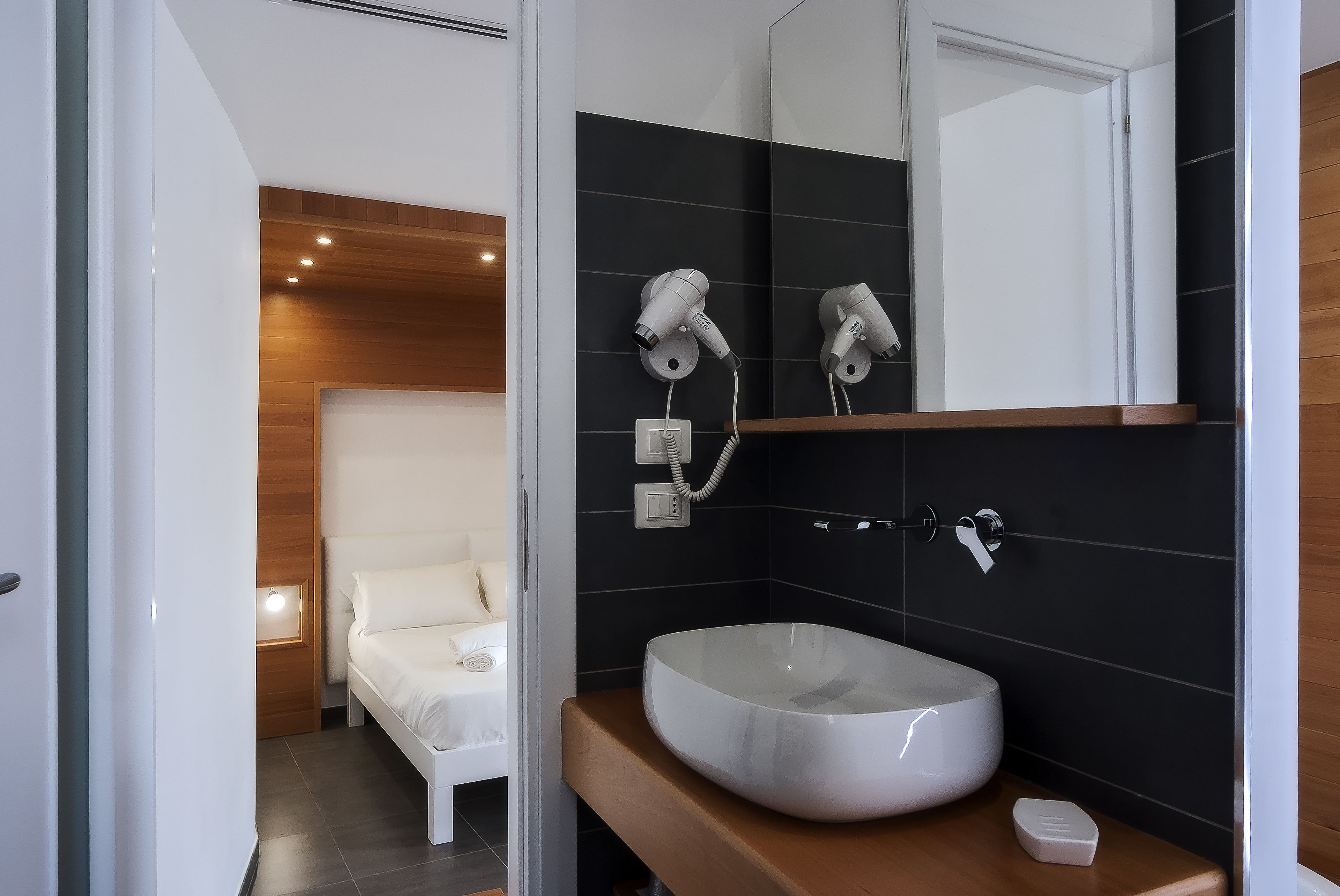 Un baño moderno con un lavamanos grande, dos secadores de pelo fijos en la pared y una mesita de noche, con una cama al fondo.