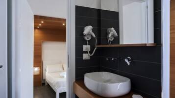 Un baño moderno con un lavamanos grande, dos secadores de pelo fijos en la pared y una mesita de noche, con una cama al fondo.