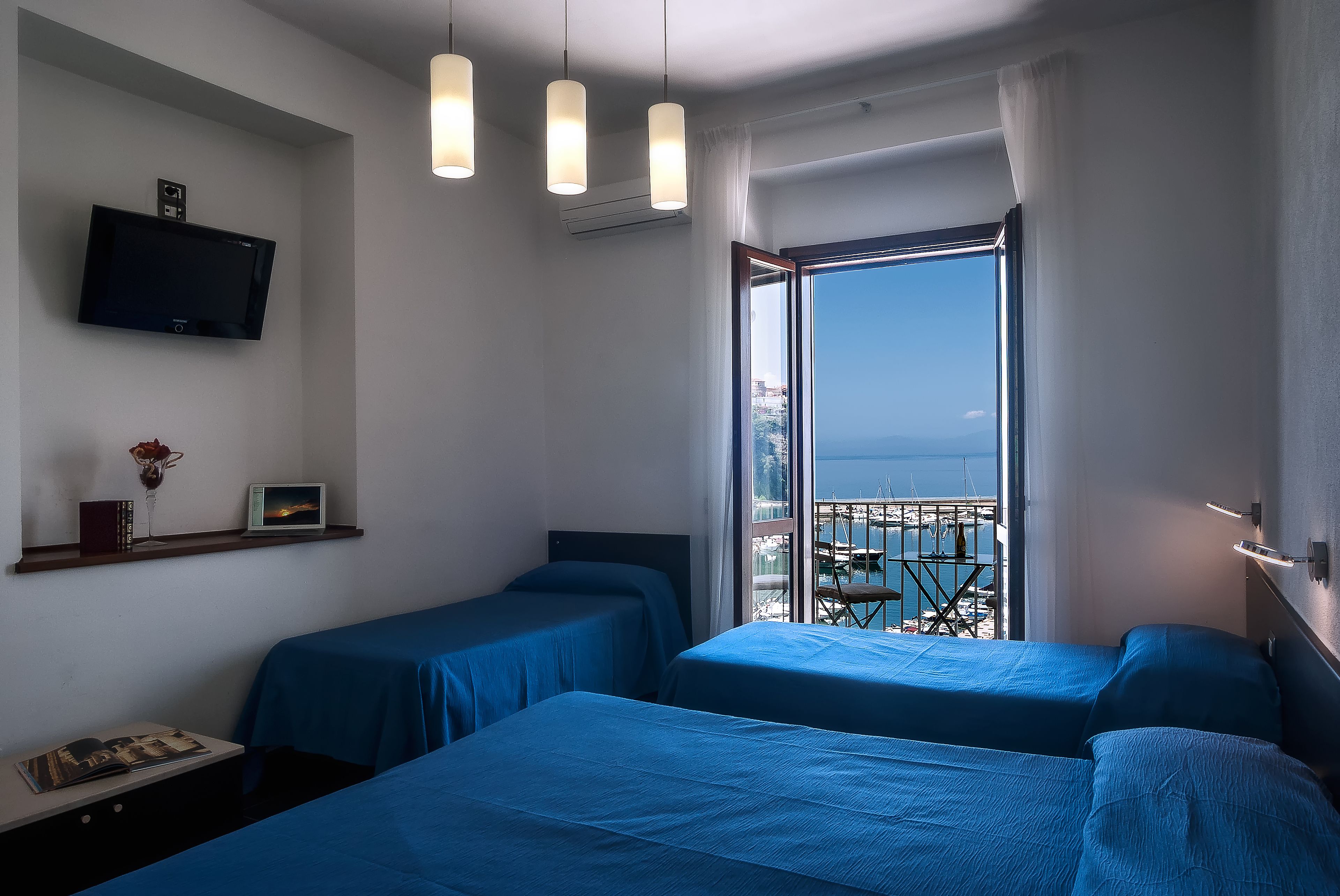 A hotel room with two beds, a TV, a shelf with books and a flower, and a balcony with a view.