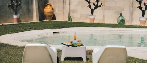 Un entorno junto a la piscina con una mesa preparada para comer, dos sillas y cactus decorativos frente a una pared de piedra.