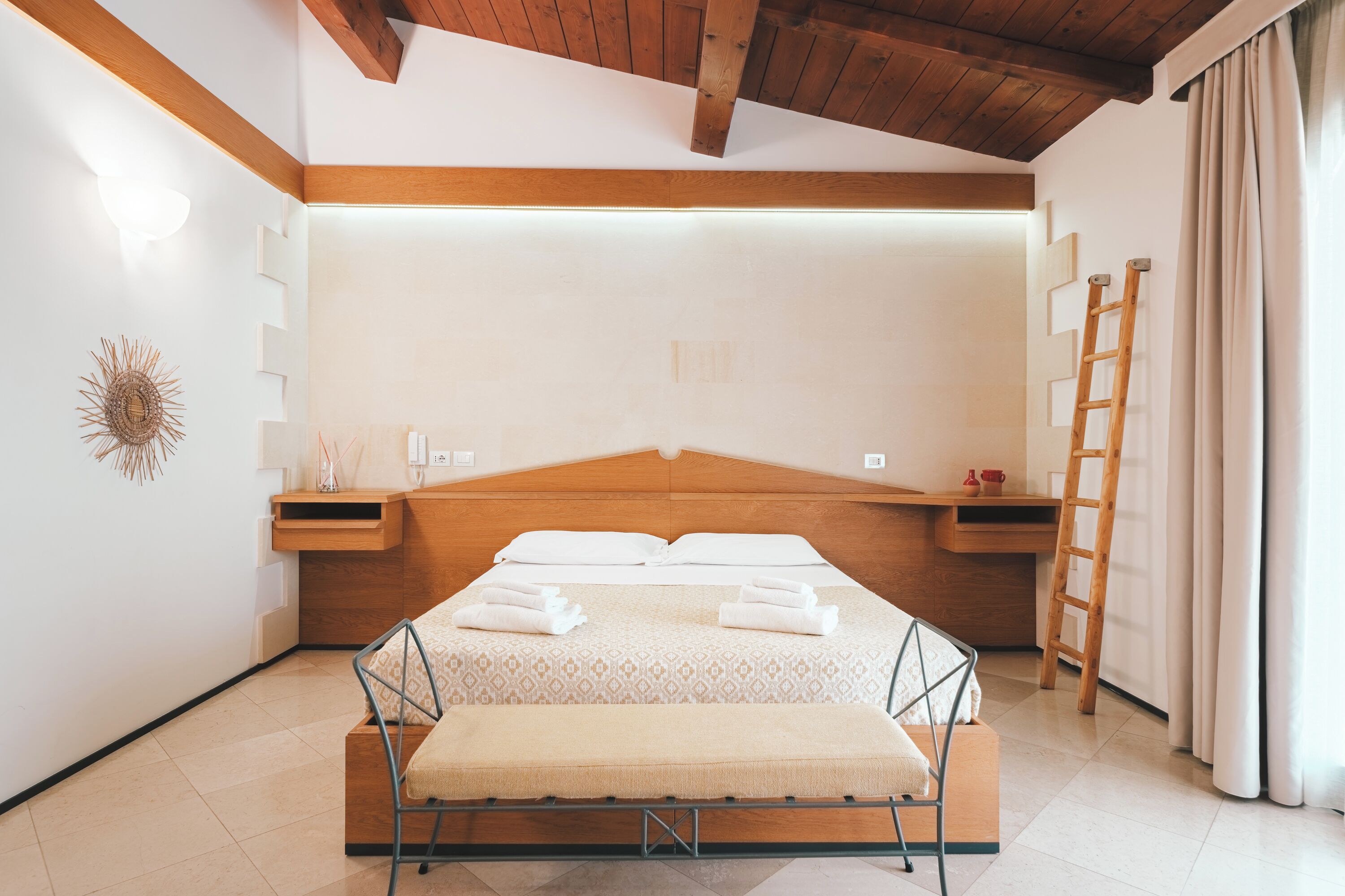 A bedroom with a wooden headboard, a bench at the foot of the bed, a ladder, and a wall-mounted shelf with a lamp and decorative items.