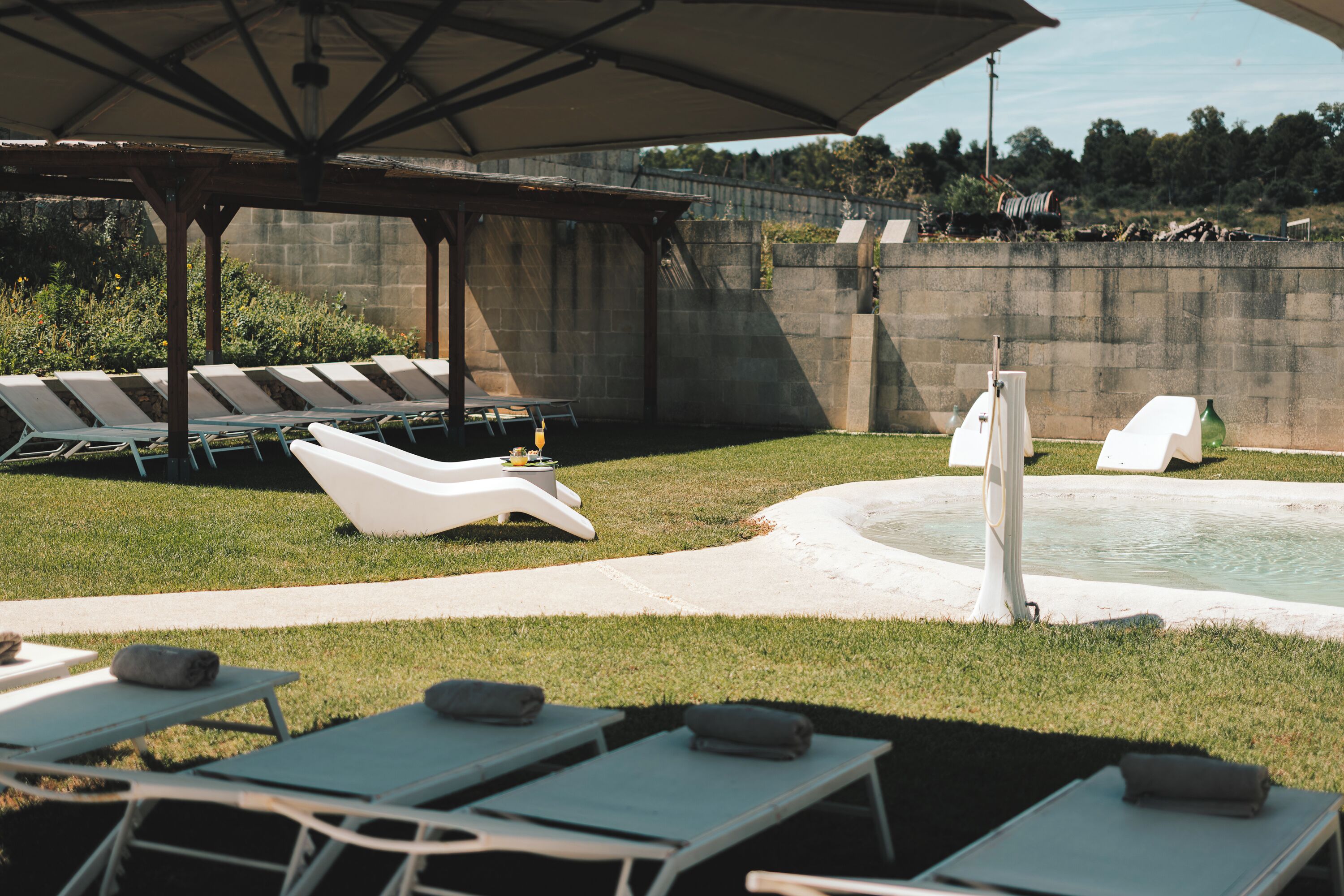 Un área junto a la piscina con sillones de descanso y sombrillas, un muro de concreto y una piscina pequeña.