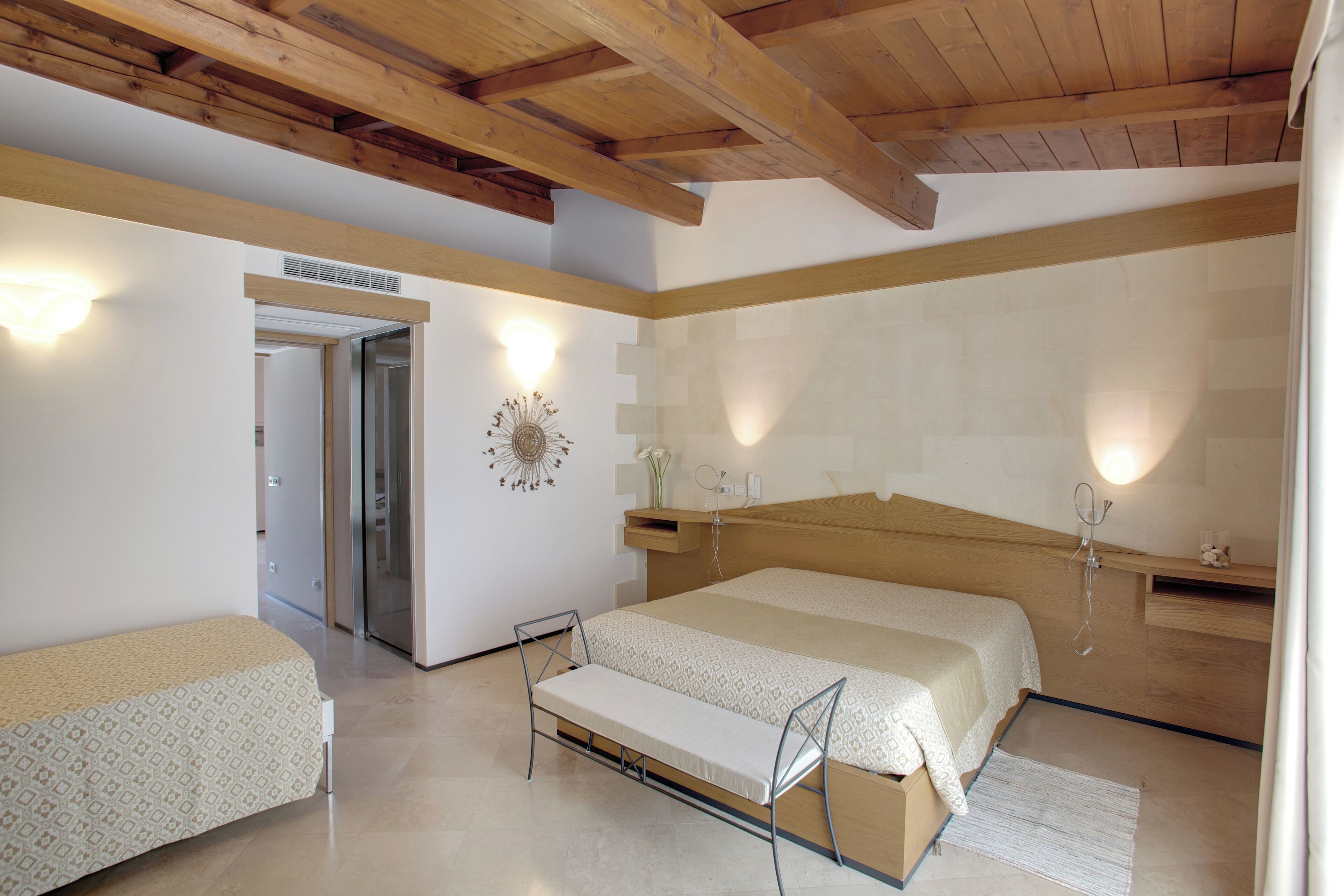 Un dormitorio con una cama grande, una cama más pequeña, un banco, vigas de madera en el techo y una lámpara fijada a la pared.