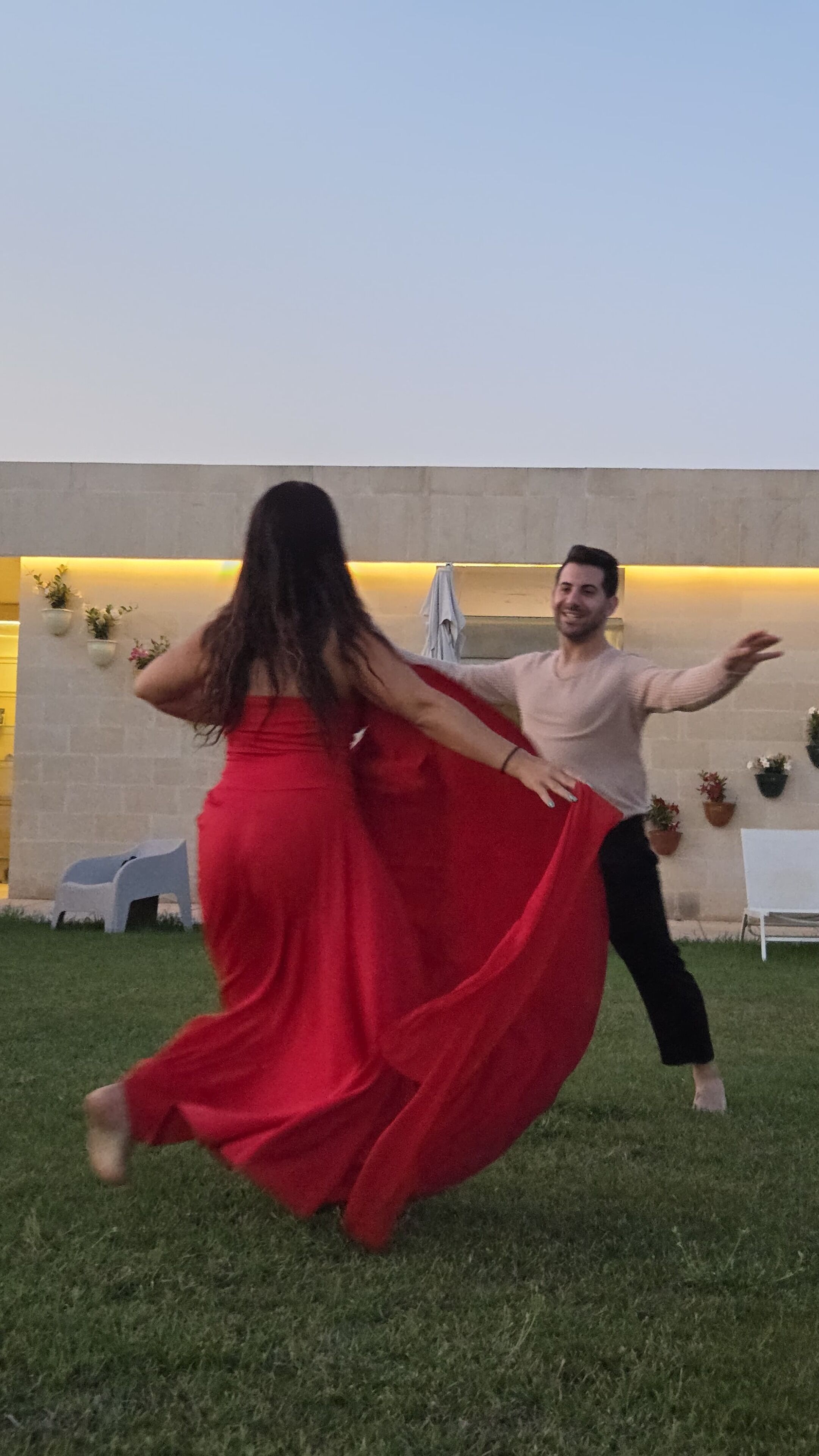 Un hombre y una mujer bailando al aire libre sobre un césped, con un muro de hormigón y plantas en macetas al fondo.