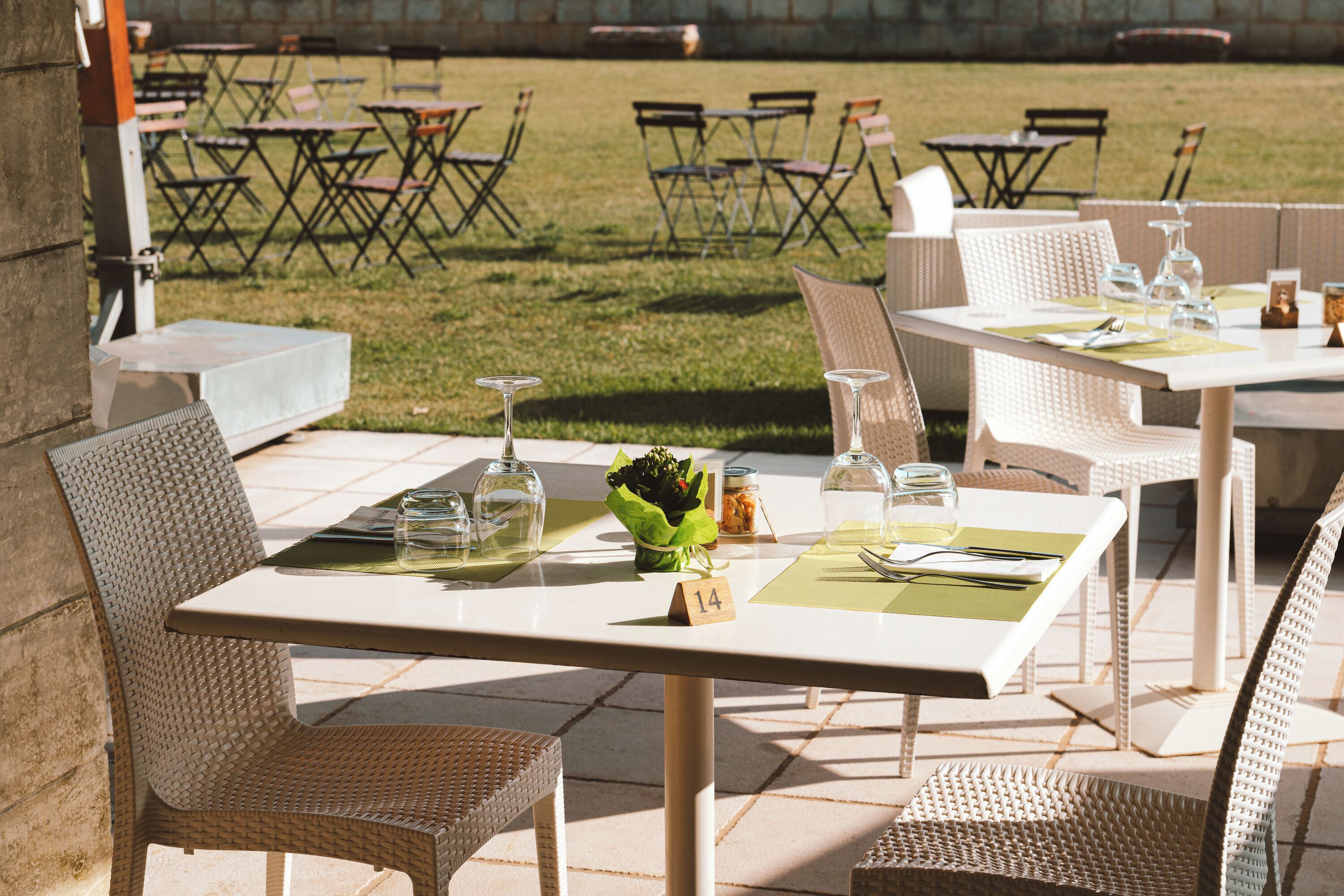 An outdoor dining area with wicker chairs and tables, some with green placemats and wine glasses.