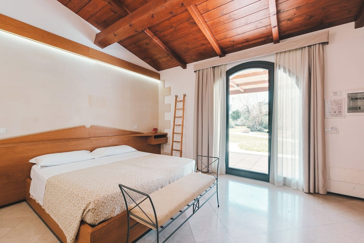 A bedroom with a wooden ceiling, a bed with a patterned bedspread, a metal ladder, and a view of the outdoors through a glass door.