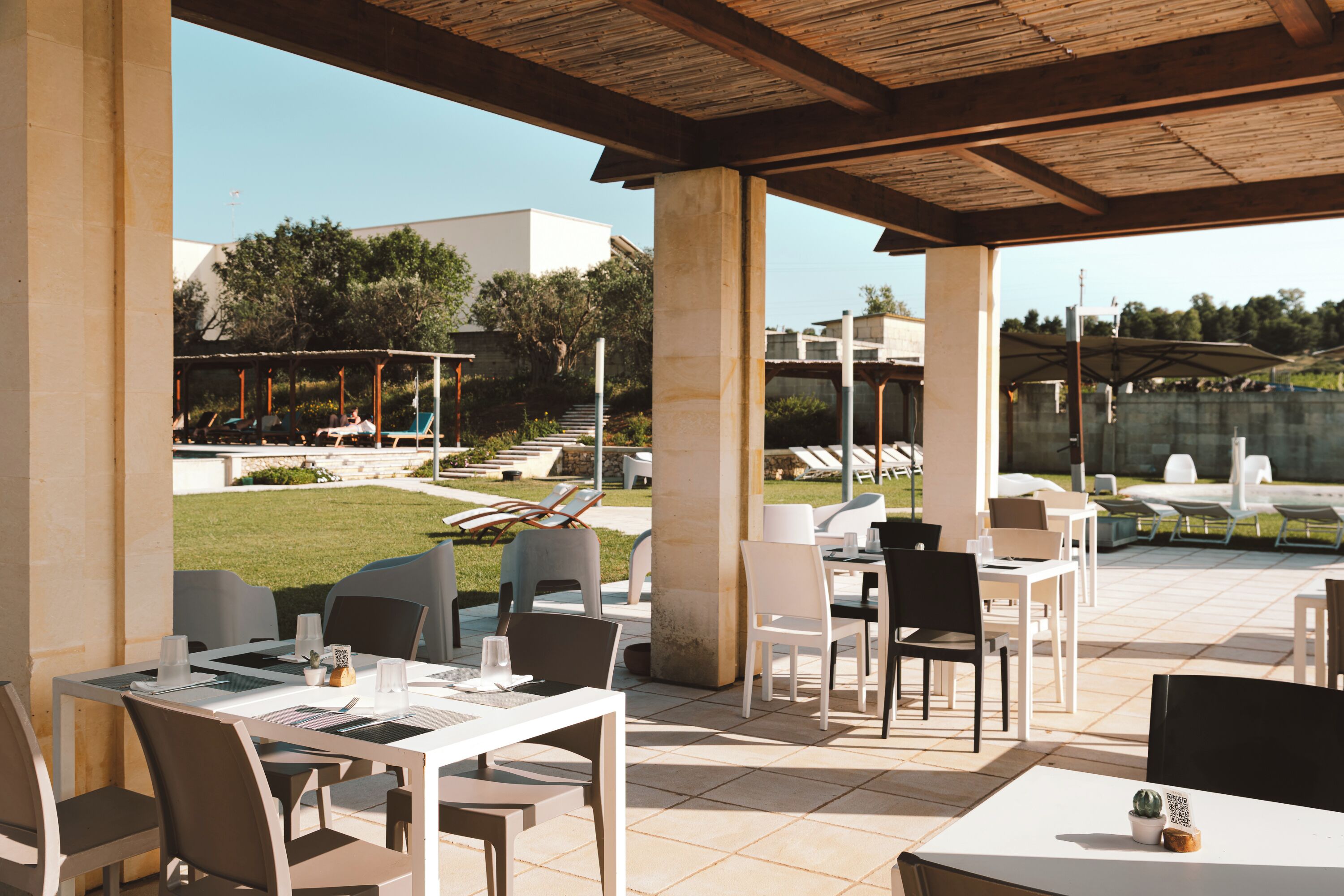 An outdoor patio area with tables and chairs, a covered seating area, and a view of a landscaped garden and buildings in the background.