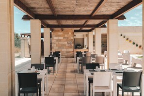 Un espace extérieur aménagé pour les repas, avec des piliers en pierre, des poutres en bois au plafond, ainsi que des tables et des chaises blanches.