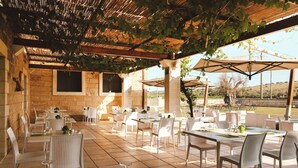 Een buiten eethoek met witte rieten stoelen en tafels, overdekt door een pergola begroeid met wijnranken.