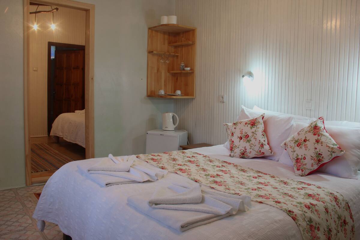 A neatly made bed with floral bedding, a bedside table with a kettle, and a wooden shelf with toiletries.