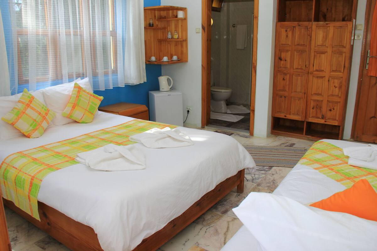 A bedroom with two beds, a wooden wardrobe, a small bathroom, and a wooden shelf with toiletries.