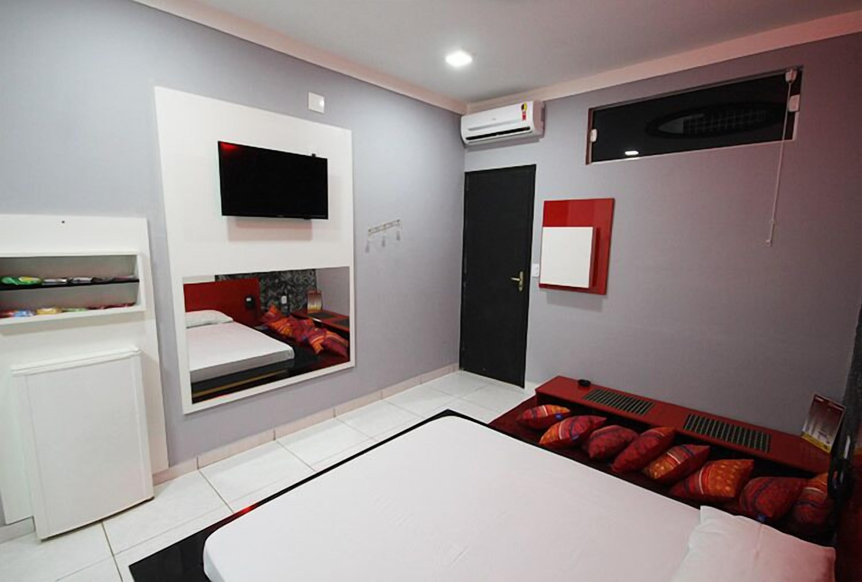 A modern hotel room with a flat-screen TV mounted on the wall, a built-in wardrobe, a bed with red and white bedding, and a small seating area with cushions.