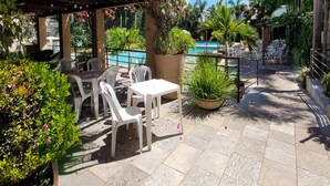 Un patio exterior con sillas y mesas de plástico blanco, rodeado de exuberante vegetación y una piscina.