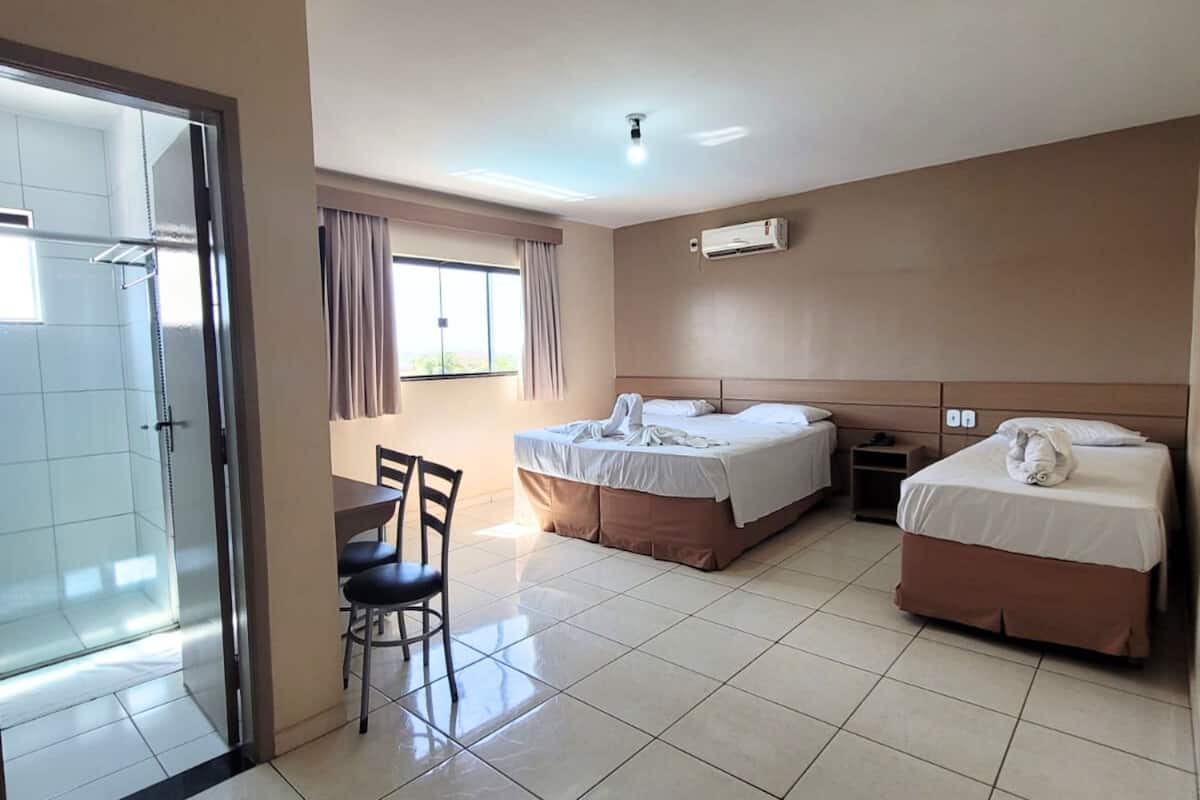 Habitación de hotel con dos camas, una mesa de comedor con sillas, un ventanal corredero y un equipo de aire acondicionado.