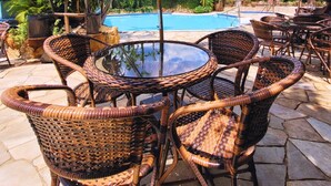 Un área junto a la piscina con sillas de mimbre y una mesa redonda bajo un paraguas grande.